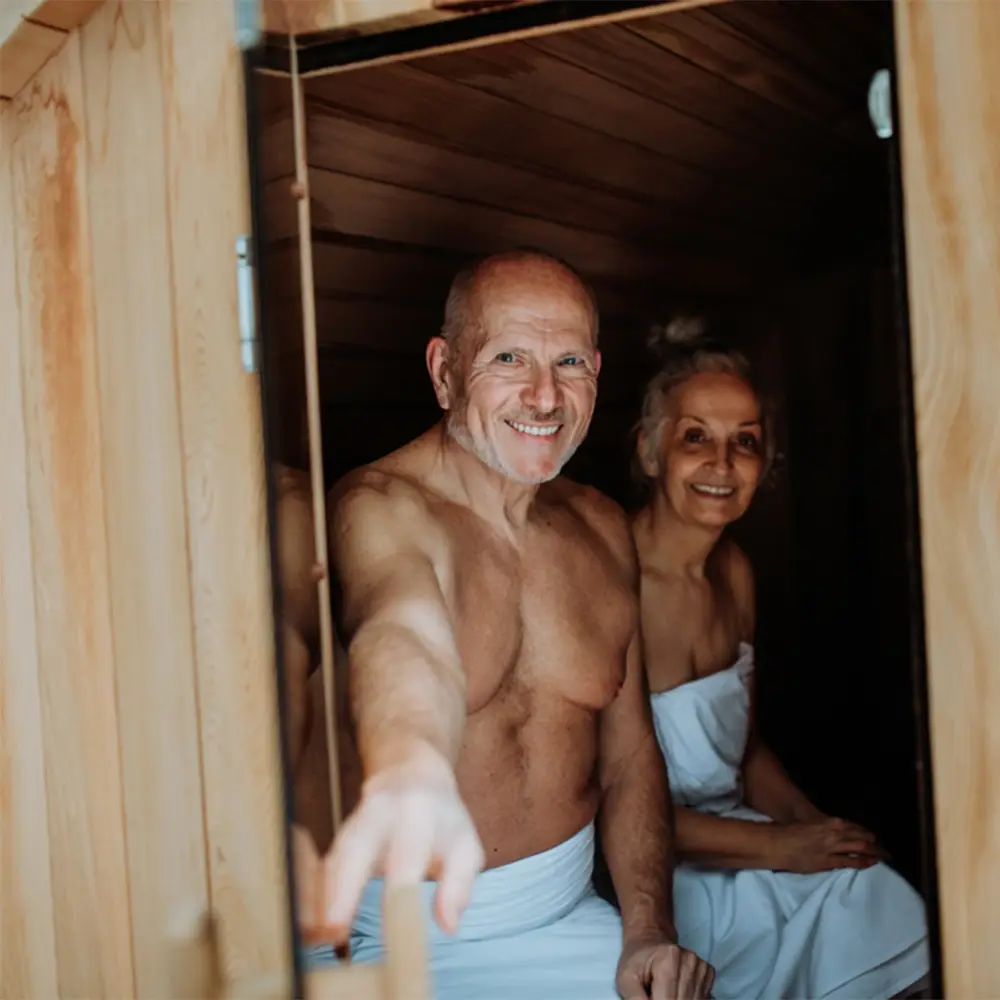 A smiling man and woman sat inside a small sauna, about the close the door